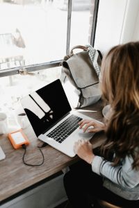 Woman working on a laptop at a wooden desk, with a backpack and coffee cup nearby, illustrating digital marketing strategies and e-commerce optimization.