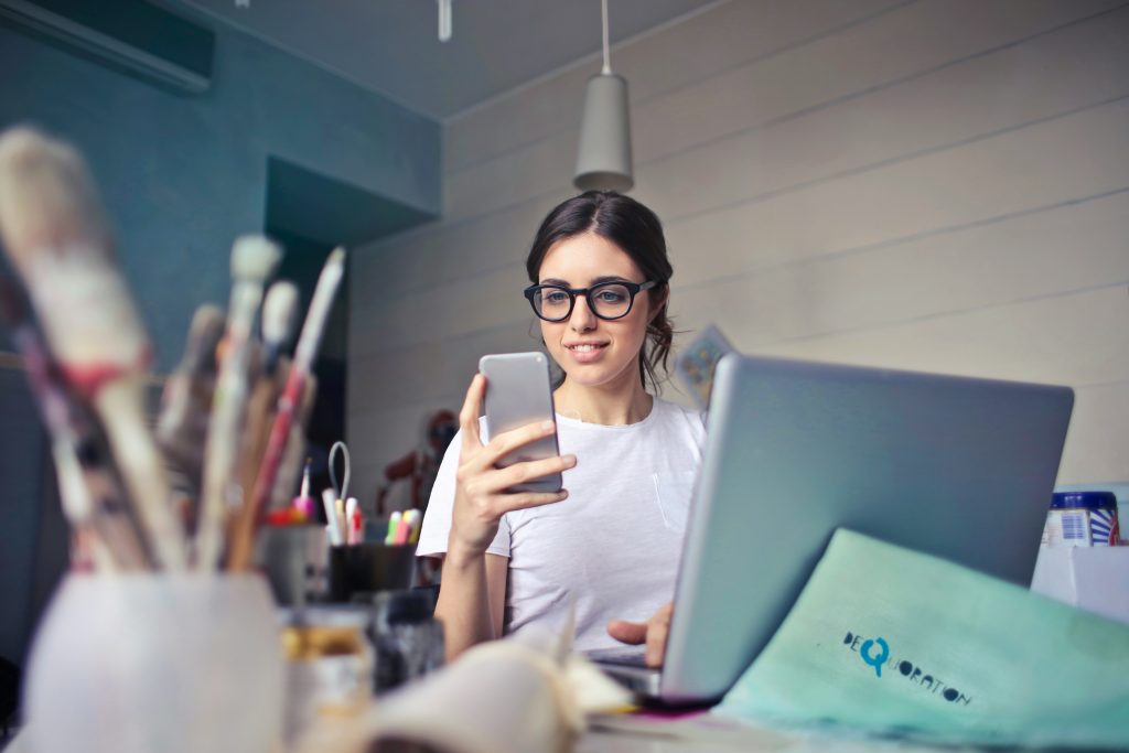 Young woman in glasses using smartphone and laptop in a creative workspace, reflecting digital marketing and content creation themes.