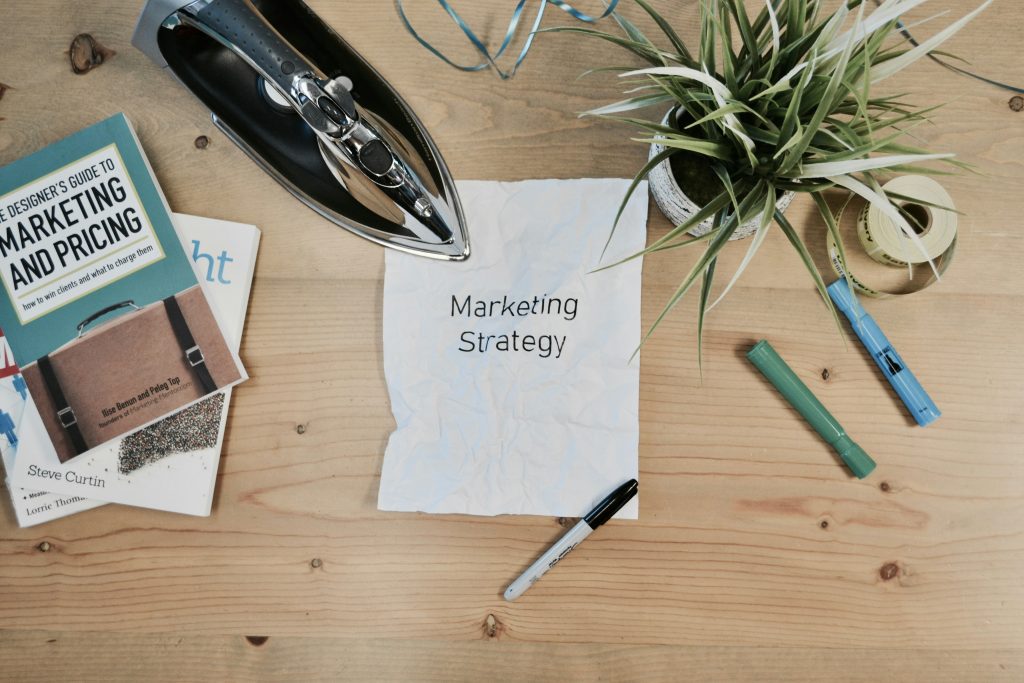 Marketing strategy document on crumpled paper surrounded by books on marketing, an iron, markers, and a small plant on a wooden table.