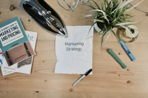 Marketing strategy document on a wooden table alongside books, pens, an iron, and a potted plant, emphasizing visual branding and effective marketing practices.