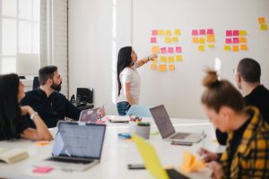 Group brainstorming session with a woman placing colorful sticky notes on a wall, surrounded by colleagues engaged in discussion and using laptops, emphasizing collaboration in digital marketing strategies.
